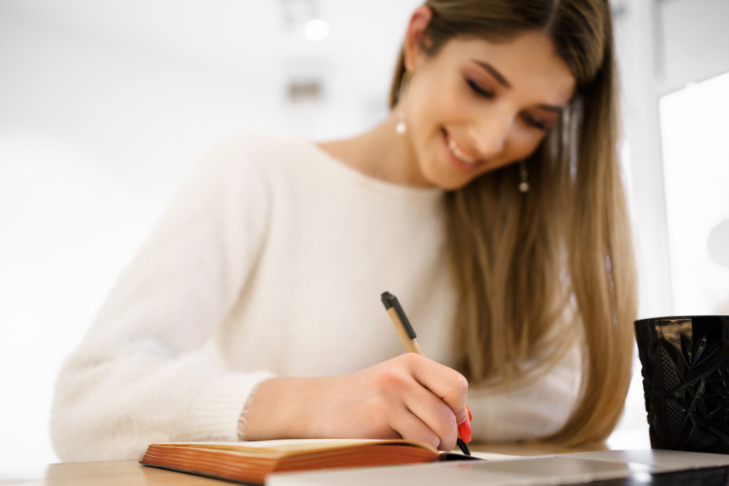 smiling-beautiful-student-female-with-long-hair-in-white-sweater-writing-in-notebook-while-using-the-laptop-remote-studing.jpg